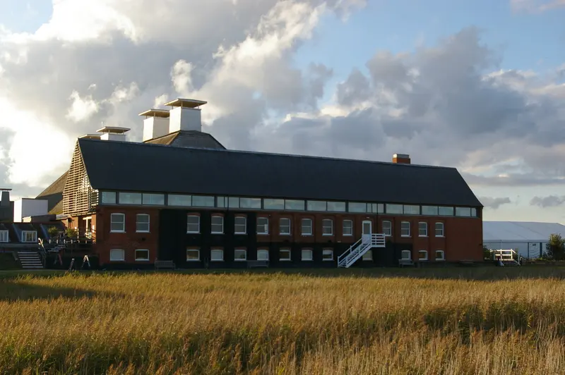 The long brick Snape Maltings concert hall with its distinctive roof vents, beyond golden reeds under a blue sky