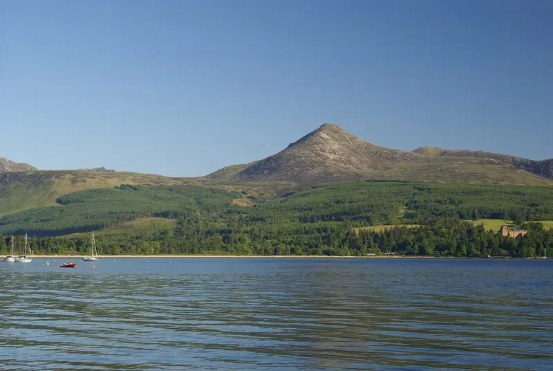The peak of Goatfell rising above wooded slopes across the calm water of Brodick Bay