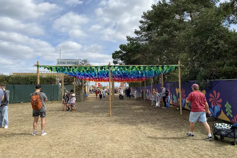A walkway under strings of colourful bunting leading into a festival site on a sunny day, with tall trees and a few visitors
