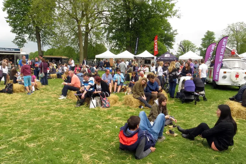 Crowds relaxing on the grass and on straw bales beneath large trees, with white food-stall gazebos behind