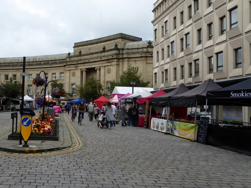 A row of black-canopied food stalls along a paved town-centre street, with flower planters and a large classical stone building behind