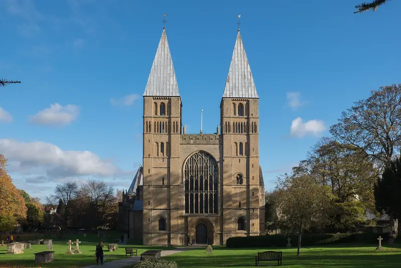 The twin pyramid-capped west towers of Southwell Minster rising above trees and lawn under a blue sky