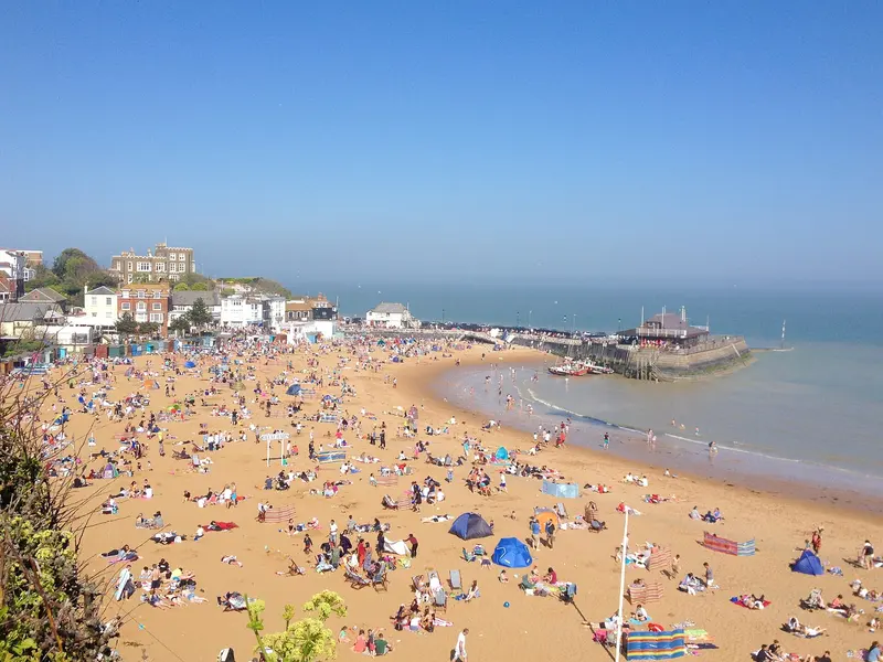 The sandy curve of Viking Bay in Broadstairs crowded with beachgoers, with the harbour pier and blue sea beyond