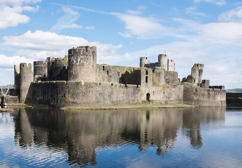 The ruined towers of Caerphilly Castle reflected in its wide moat under a blue sky