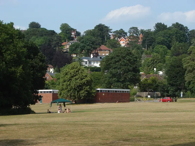 The open grass of Shalford Park with trees and wooded hillside houses beyond