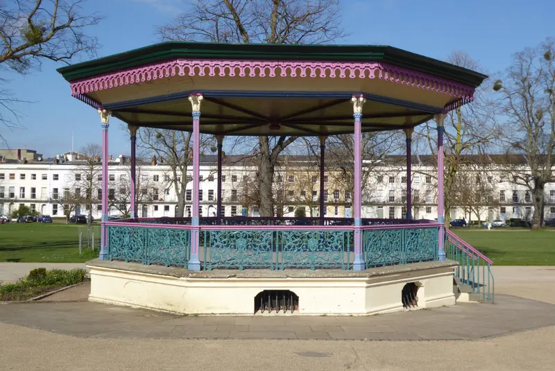 An ornate Victorian bandstand with a striped canopy in a park, with Regency terraces behind under a blue sky