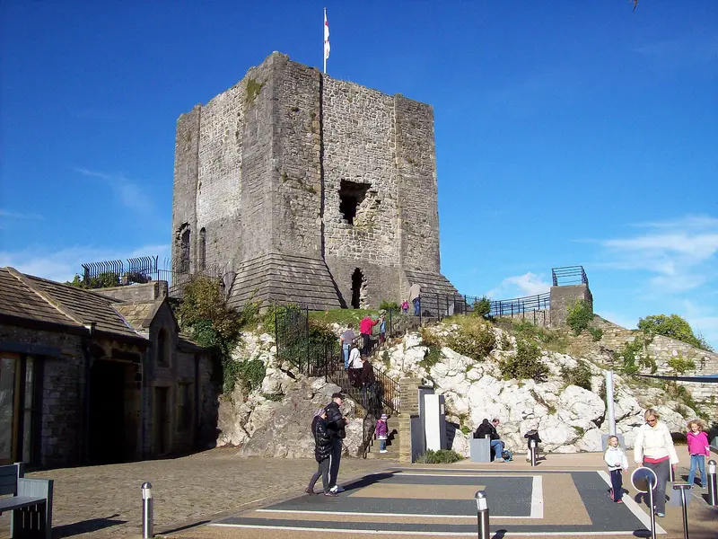 The small stone keep of Clitheroe Castle on its limestone outcrop under a deep blue sky, with visitors on the path below