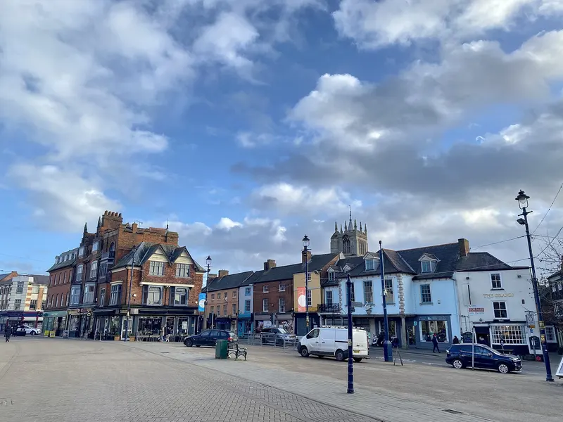 Melton Mowbray's Market Place with brick and rendered shopfronts and a church tower behind, under a blue sky with clouds