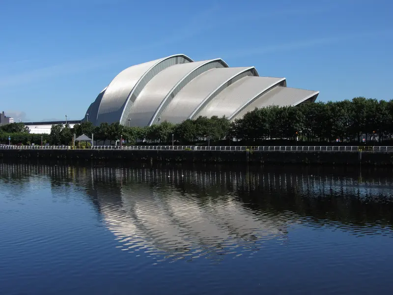 The curved, shell-like roof of the SEC Armadillo reflected in the calm River Clyde under a blue sky