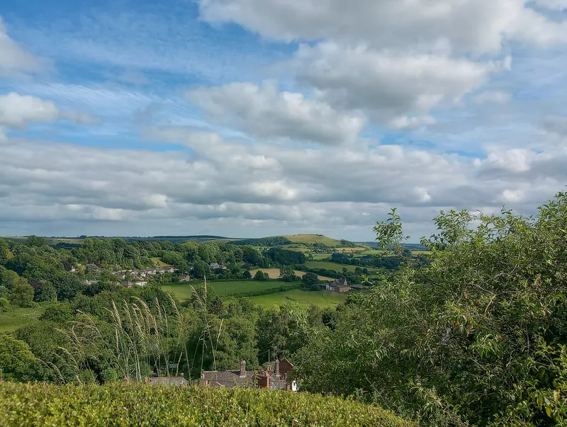 A wide view from a hilltop over green, wooded countryside under a cloudy blue sky