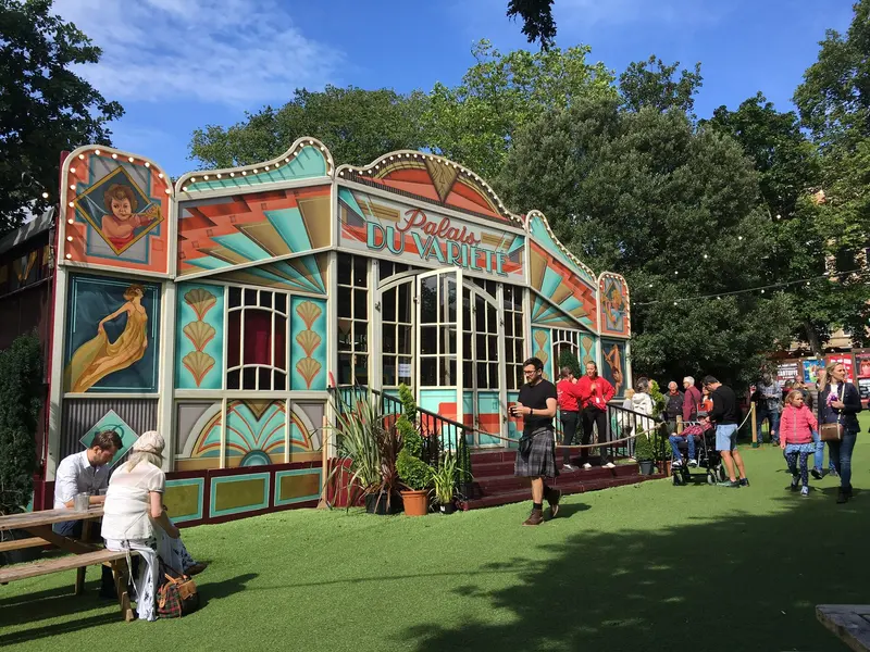 A colourful painted spiegeltent among trees in George Square Gardens, with people sitting on the grass outside