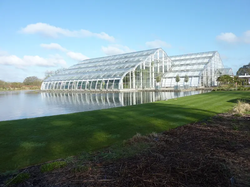The modern angular Glasshouse at RHS Garden Wisley reflected in a lake, beyond a sloping lawn