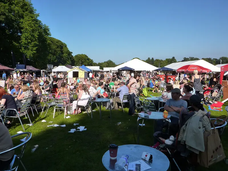 Visitors sitting at tables on the grass in sunshine, with marquees, food stalls and trees behind