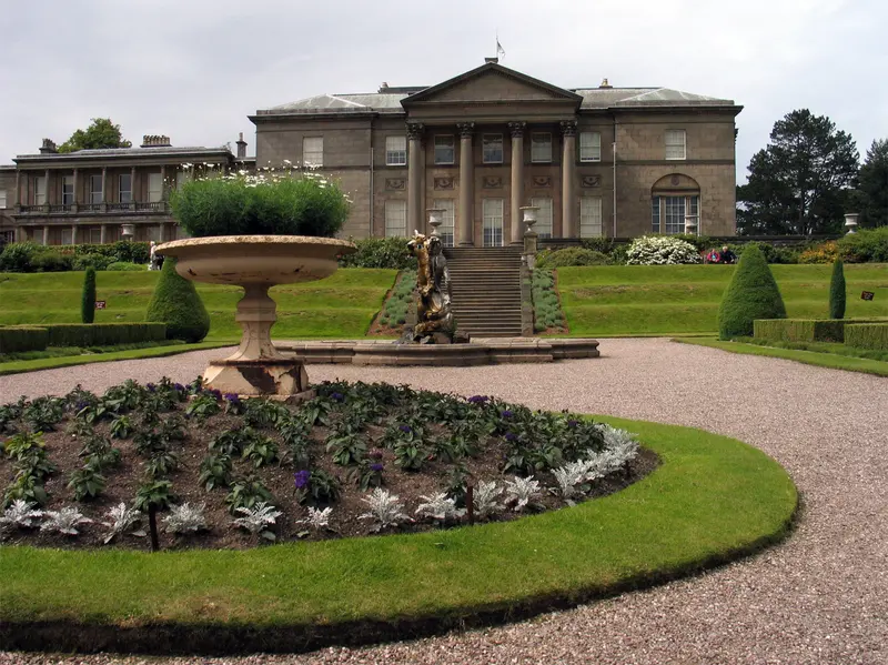 The pale stone neoclassical front of Tatton Hall above formal gardens with a fountain and flower beds