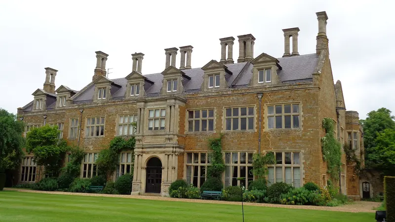 The gabled honey-coloured stone front of Holdenby House behind a lawn and flower borders