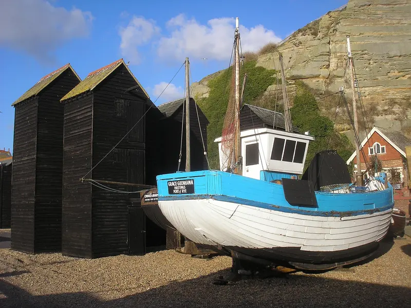 Tall black weatherboarded net shops and a blue fishing boat on the shingle beach of the Stade in Hastings