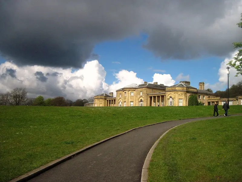 The pale neoclassical Heaton Hall beyond a curving path and lawn, under dark clouds with patches of blue sky