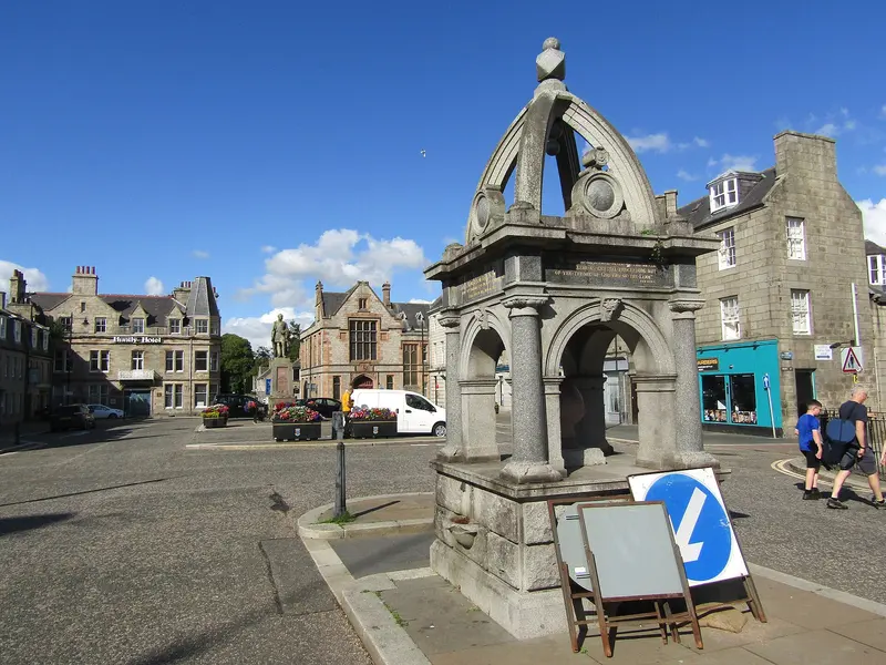 A Victorian stone drinking fountain in the middle of Huntly's town square, surrounded by granite buildings under a blue sky