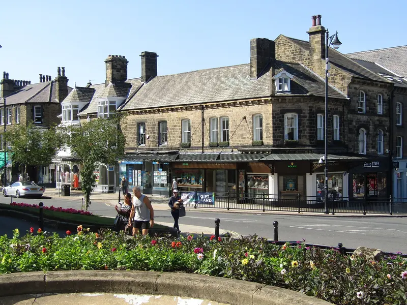 Stone Victorian buildings and shops in Ilkley town centre, with flower beds in the foreground under a blue sky