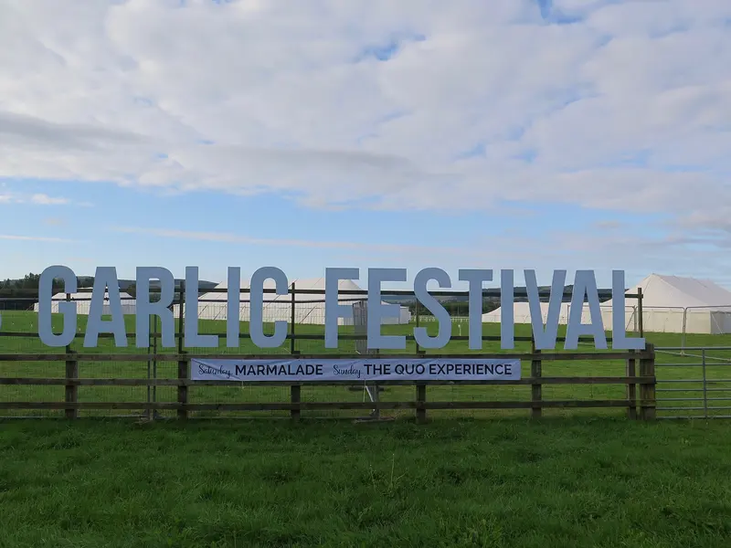 Large 'Garlic Festival' letters standing on a grassy field, with white marquees and a wide cloudy sky behind