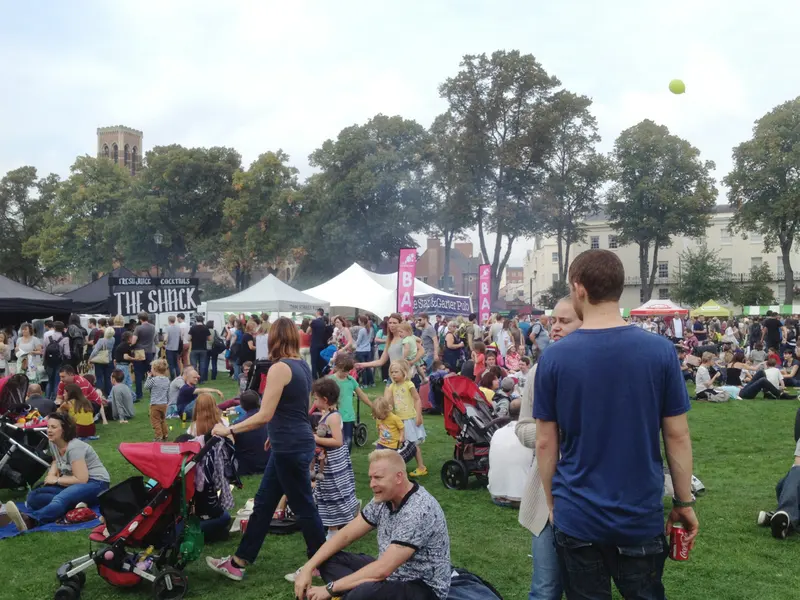 Crowds of visitors on the lawns among festival stalls and gazebos in a town park, with trees and buildings behind