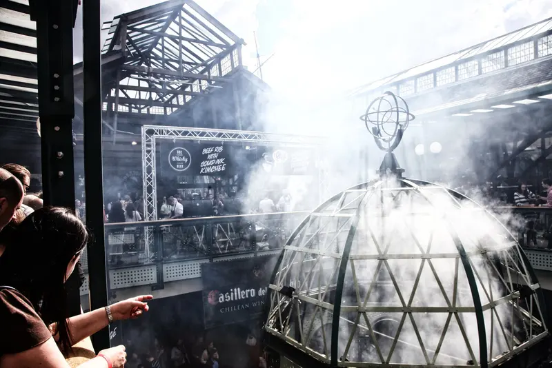 Smoke rising from a glass-domed grill inside the iron-framed warehouse hall of Tobacco Dock, with festival-goers nearby