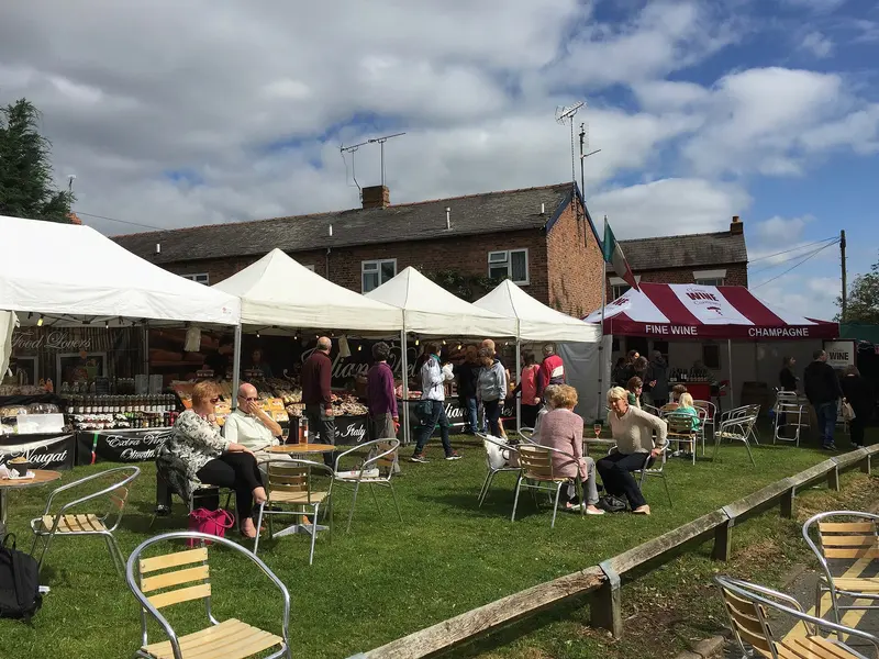 Visitors at tables on a lawn beside a row of white festival marquees, with brick town buildings and a blue sky behind
