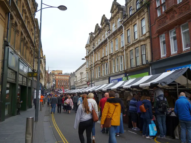 Shoppers walking between rows of canopied food stalls along a city-centre street lined with Victorian buildings
