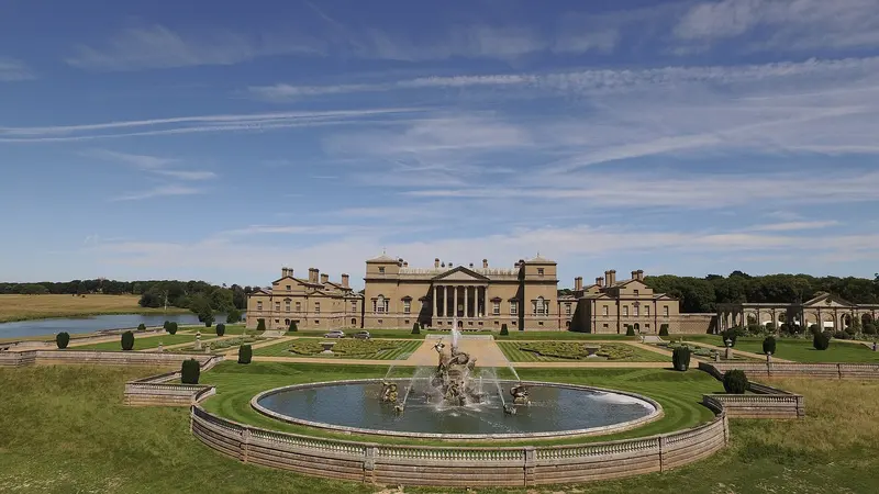 The long Palladian south front of Holkham Hall beyond a lawn and a large fountain under a blue sky