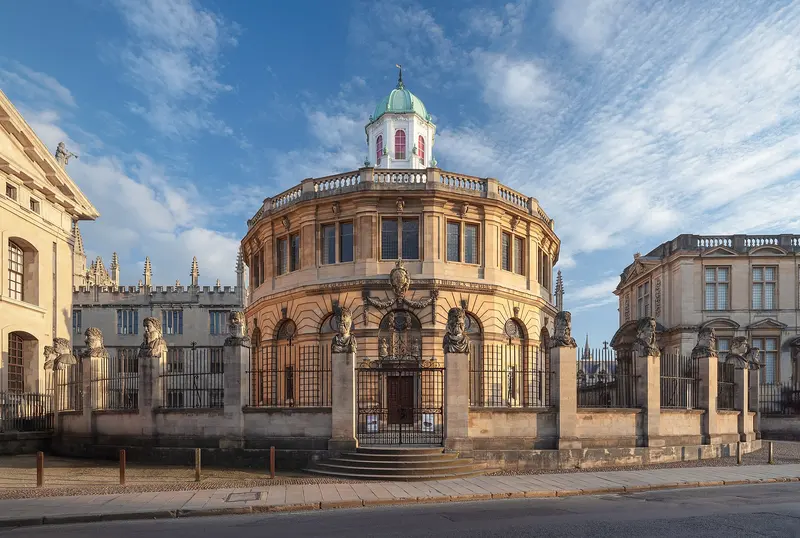 The domed, round Sheldonian Theatre behind its railings and stone heads on Broad Street, Oxford, under a blue sky