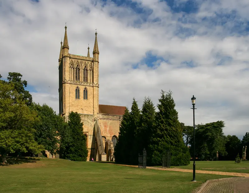 The tall Gothic tower of Pershore Abbey beside trees and a lawn under a cloudy blue sky