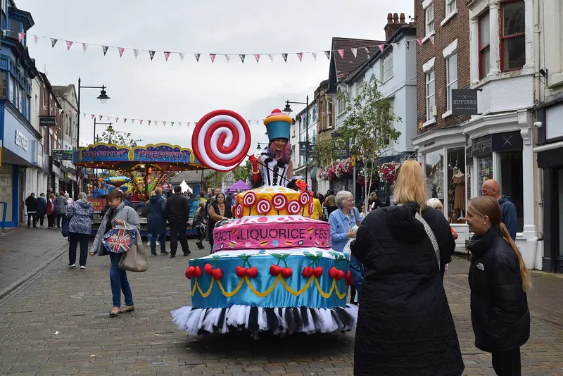 A sweet-themed parade float shaped like a giant tiered cake, with a costumed performer on top, moving along a bunting-hung town street as onlookers watch