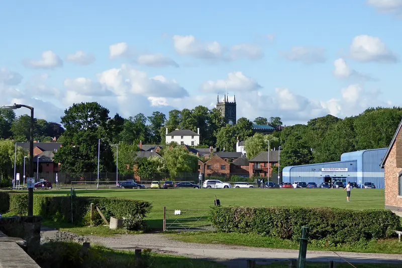The open lawns of Westbridge Park in Stone, with the town and a church tower beyond under a blue sky