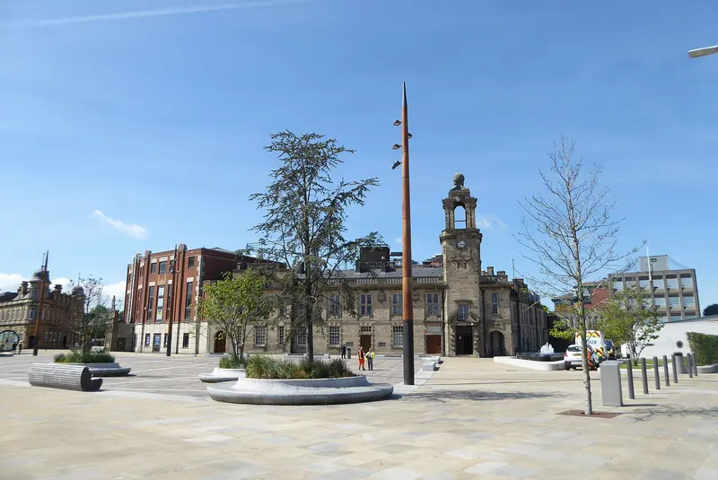 The open paved Keel Square in Sunderland city centre, with young trees, a tall mast-like sculpture and surrounding buildings under a blue sky