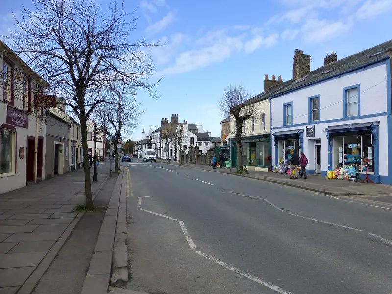 A wide, tree-lined Main Street in Cockermouth with colourful painted shopfronts under a blue sky