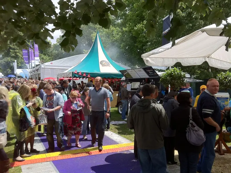 Crowds of visitors walking along a coloured walkway between white marquees and stalls under trees