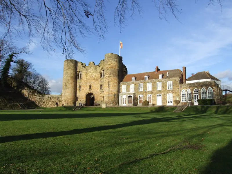 The twin-towered stone gatehouse of Tonbridge Castle beside a Georgian house, above a lawn under a blue sky