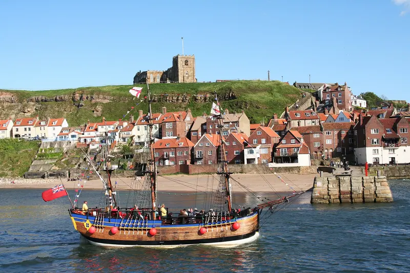 A replica sailing ship in Whitby harbour, with red-roofed houses climbing the East Cliff towards St Mary's Church