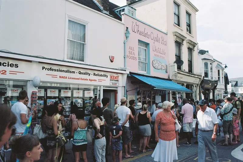A crowd of people gathered along a narrow seaside high street lined with shops