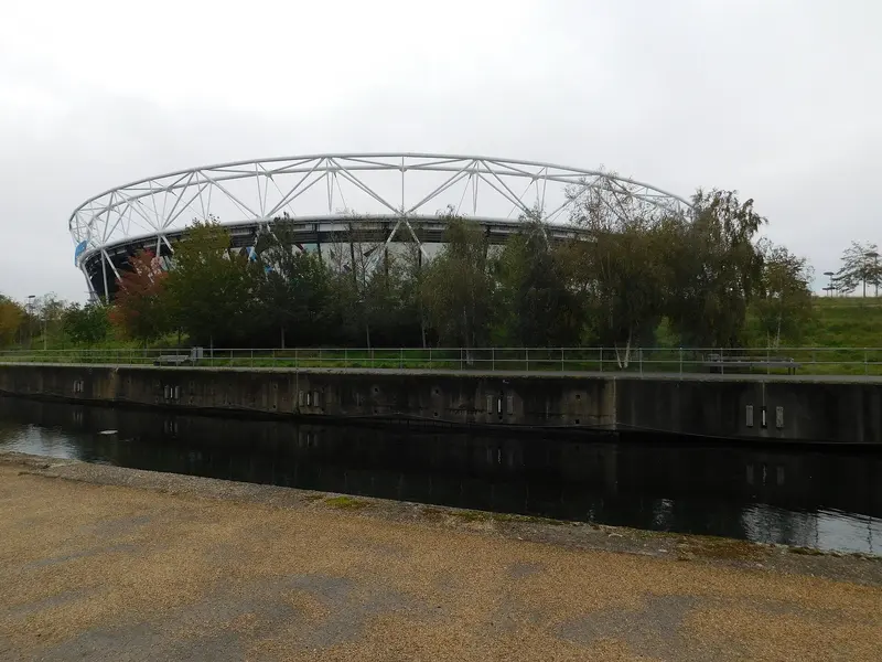 The London Stadium's white roof structure seen across a river and trees in Queen Elizabeth Olympic Park