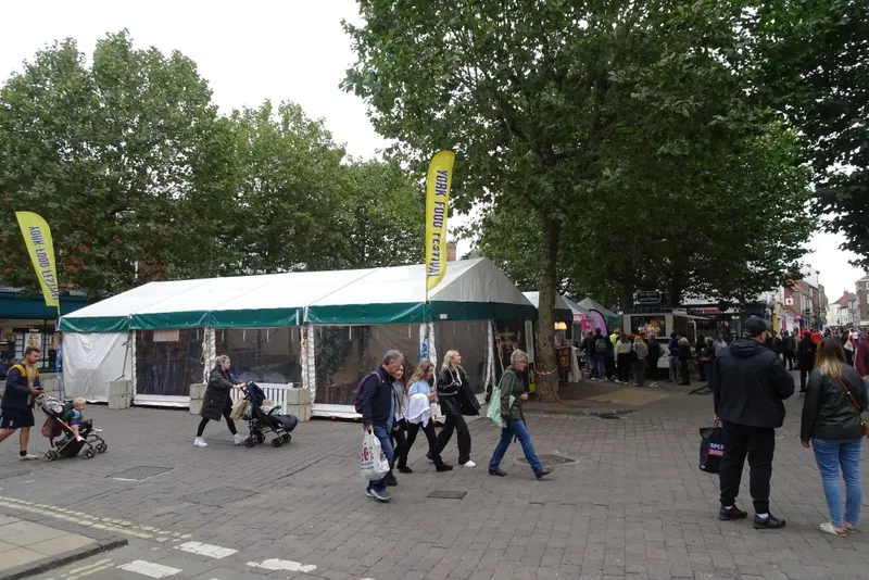 Festival marquees and food stalls under trees in a paved city square, with visitors walking past