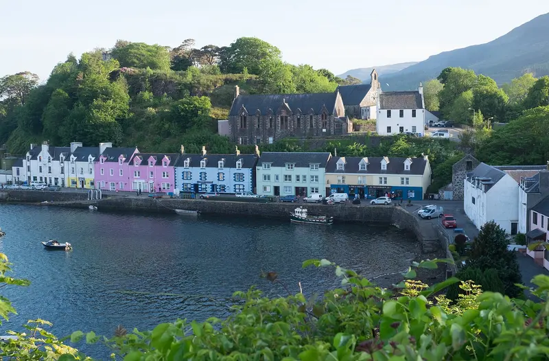 A row of brightly painted houses along Portree harbour on the Isle of Skye, with boats in the bay and wooded hillside above